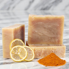 Three rectangular bars of handmade Turmeric Lemon Exfoliating Shea Butter soap in a warm peach-beige tone are arranged on a white marble surface, with lemon slices and a small pile of Turmeric powder in front by Adiva Naturals 