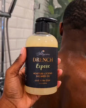 A hand holds a bottle of Drench Expose Moisture-Locking Shower Oil in front of a person in a shower, with a tiled bathroom background and shower head visible.