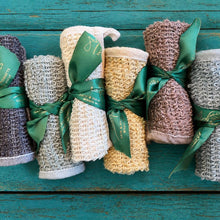 Six rolled bath towels in neutral shades of gray, beige, cream, and brown, tied with green satin ribbons, arranged vertically on a distressed teal wooden background. The ribbons feature the brand name 'Bath & Natural'.