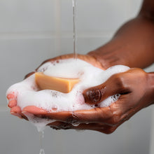 A close-up of a person's hands washing under running water, holding a bar of Turmeric Lemon Exfoliating Soap covered in white lather foam.