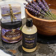 Lavender-themed beauty products including a bottle of Drench Tranquility shower oil, a jar of Total Body Exfoliation Lavender, and a wrapped lavender soap bar on a stone surface with a wooden bowl of lavender flowers.