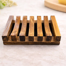 A rectangular wooden soap dish with six parallel raised slats, shown on a light stone surface with softly blurred decorative props in the background.