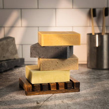 Four rectangular bars of soap in different earthy tones stacked on a wooden soap dish, set on a countertop with a tiled wall and metal container in the background.